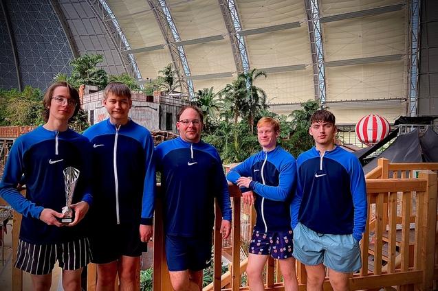 Fünf Personen in blauen Sportjacken stehen vor einer grünen, tropischen Kulisse und halten einen Pokal.