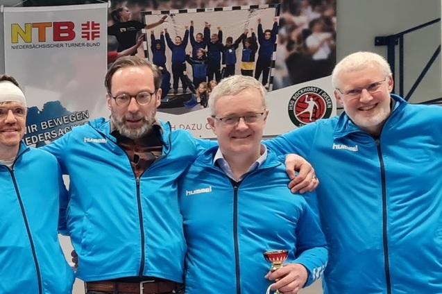 Vier Männer in blauen Trainingsjacken stehen lächelnd zusammen, ein Mann hält einen Pokal. Im Hintergrund ist ein Banner.