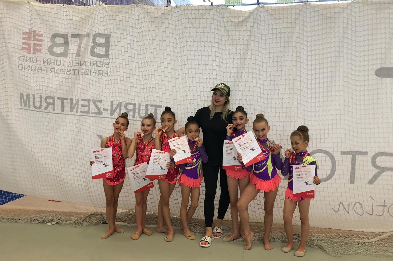 Sieben junge Turnerinnen in bunten Kostümen zeigen ihre Urkunden und Medaillen neben einer Trainerin.