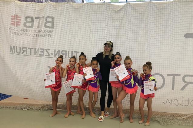 Sieben junge Turnerinnen in bunten Kostümen zeigen ihre Urkunden und Medaillen neben einer Trainerin.