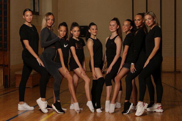 Gruppe von neun Tänzerinnen in Sportkleidung posiert auf einem Holzfußboden in einer Turnhalle.