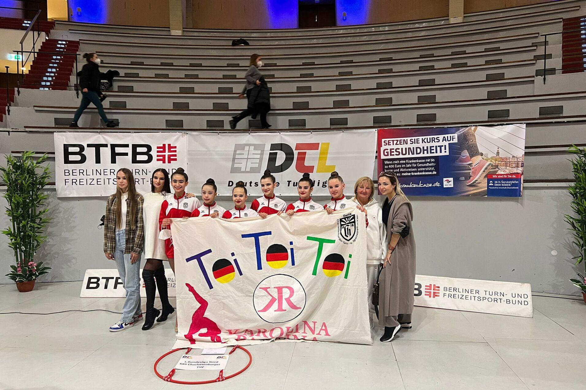Gruppenshot mit sechs Tänzerinnen in roten Outfits und zwei Frauen, die ein Banner mit „Toi Toi Toi“ halten.