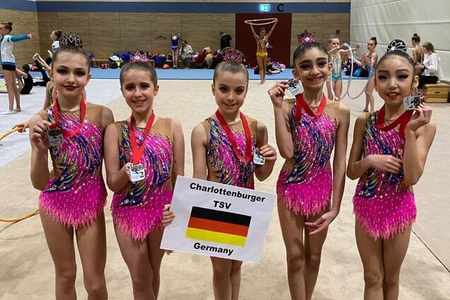 Fünf junge Mädchen in bunten Gymnastikanzügen halten Medaillen und ein Schild mit „Charlottenburger TSV, Germany“.