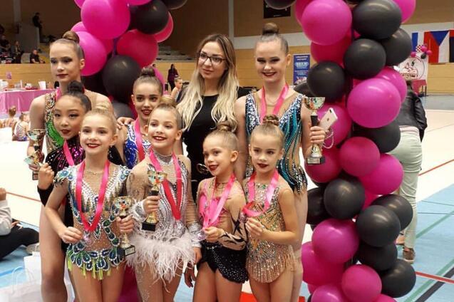 Gruppe von Gymnastinnen in bunten Kostümen mit Pokalen und Medaillen vor einem Luftballonbogen in einer Sporthalle.