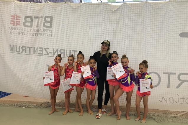 Sieben junge Turnerinnen in bunten Kostümen zeigen ihre Urkunden und Medaillen neben einer Trainerin.