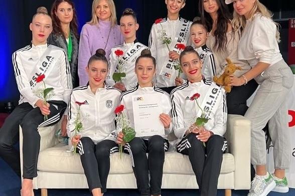 Gruppe von Tänzerinnen in schwarz-weißen Trainingsanzügen, die mit Urkunde und Rosen auf einem Sofa posieren.