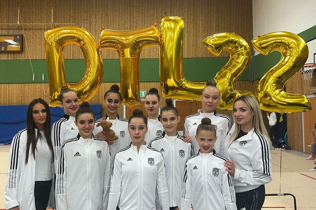 Gruppe von Tänzerinnen in weißen Trainingsjacken, posierend vor großen goldenen Ballons mit der Aufschrift „DTL22“.