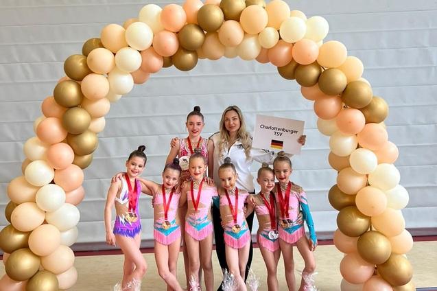 Gruppe junger Turnerinnen in bunten Kostümen mit Medaillen, umgeben von Luftballons, posiert mit ihrer Trainerin.