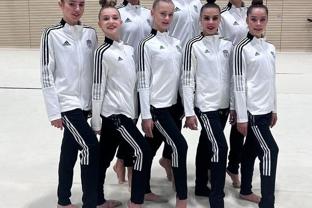 Gruppe von acht sportlichen Mädchen in weißen Trainingsjacken und schwarzen Hosen, posierend auf einem hellen Sportboden.