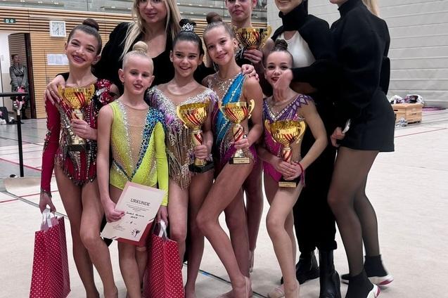 Gruppenszene mit erfolgreichen Gymnastinnen, die Pokale und Urkunden präsentieren, umgeben von Trainerinnen in einem Wettkampf.