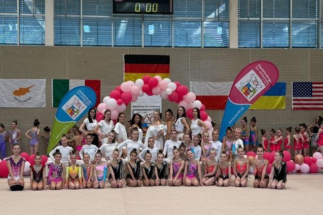 Gruppenfoto von Gymnastinnen und Trainerinnen vor dekorierten Wänden mit Nationalflaggen und einem Luftballonbogen.