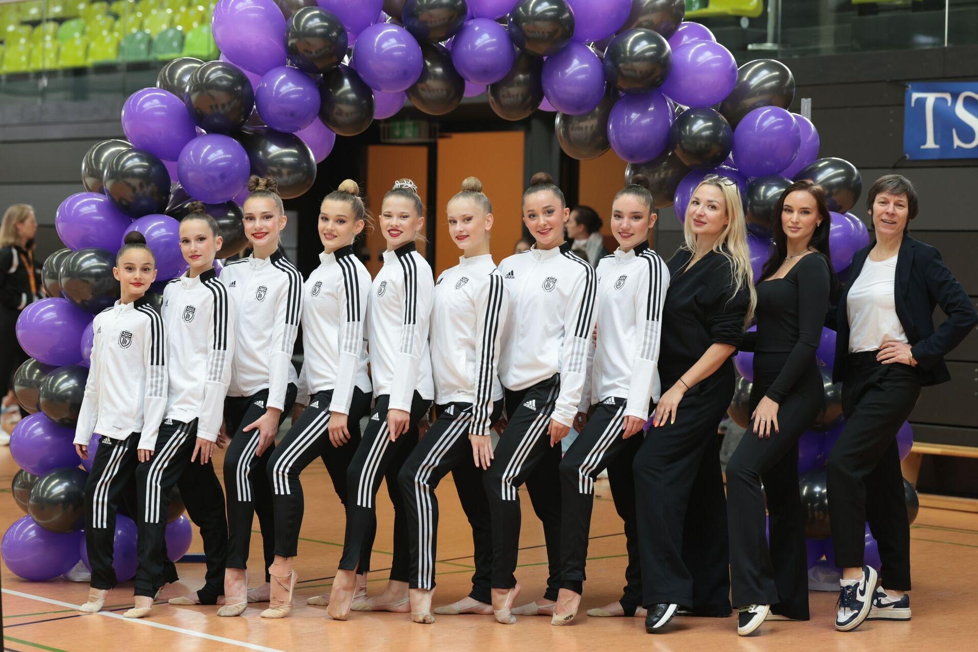 Gruppe von Tänzerinnen in Trainingsanzügen vor einem Luftballonbogen in Lila und Schwarz in einer Sporthalle.