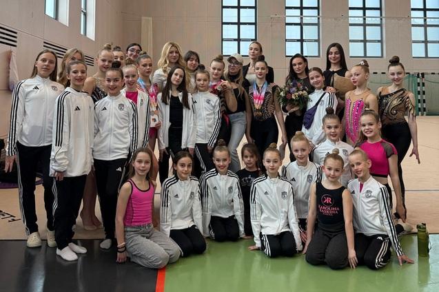 Gruppenfoto von Gymnastinnen in Trainingsanzügen, umgeben von ihren Trainerinnen in einer Sporthalle.