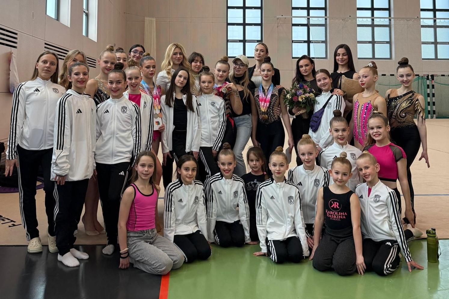 Gruppenfoto von Gymnastinnen in Trainingsanzügen, umgeben von ihren Trainerinnen in einer Sporthalle.