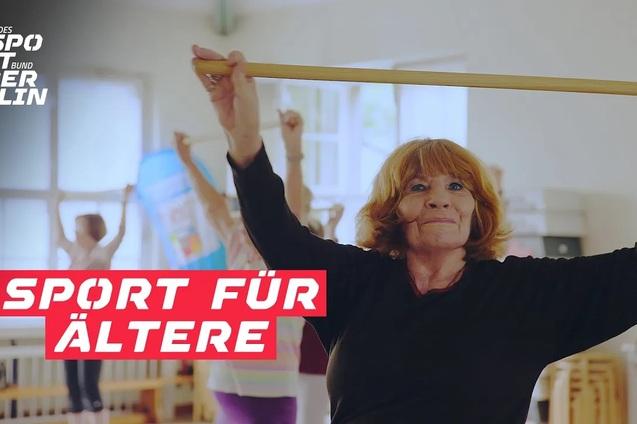 Ältere Frau hebt einen Stab in einem Sportkurs, im Hintergrund sind weitere aktive Teilnehmer zu sehen.