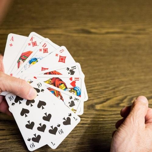 Nahaufnahme von zehn Spielkarten in einer Hand, darunter Asse und Figuren, auf einem Holz-Tisch.