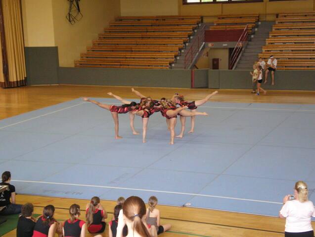 Sechs Turnerinnen in roten und schwarzen Einheitenführen eine synchronisierte Übung auf der blauen Turnmatte vor.