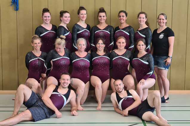 Gruppe von 14 Sportlern in bordeauxfarbenen Trikots mit Glitzer, posing in einer Turnhalle.
