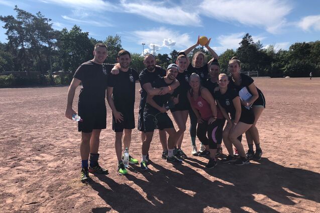Gruppe von zehn Sportlern in schwarzen Trikots posiert auf einem Sportplatz mit blauem Himmel im Hintergrund.