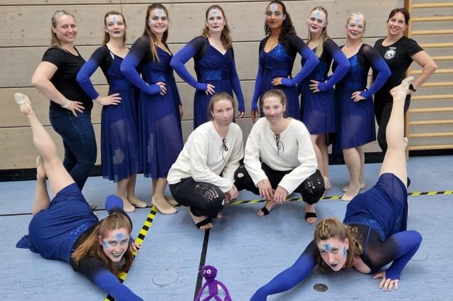 Gruppe von Tänzerinnen in blauen Kostümen posiert auf einer Sportfläche, einige in akrobatischen Posen, mit geschminkten Gesichtern.