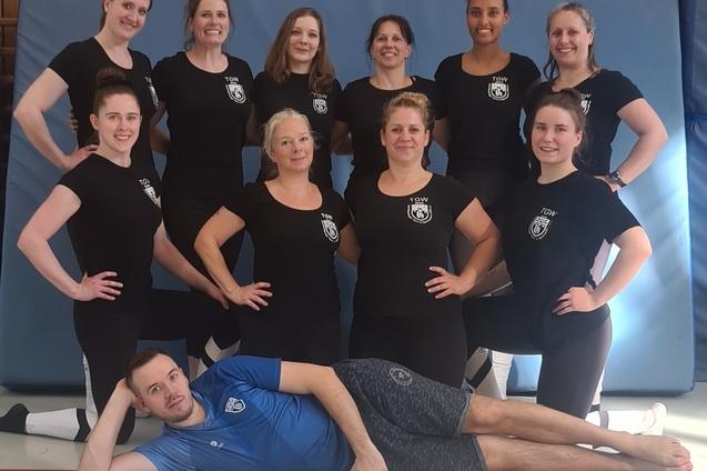 Gruppenschnappschuss von zehn Frauen und einem Mann in Sportkleidung, posierend vor einer Turnmatte.