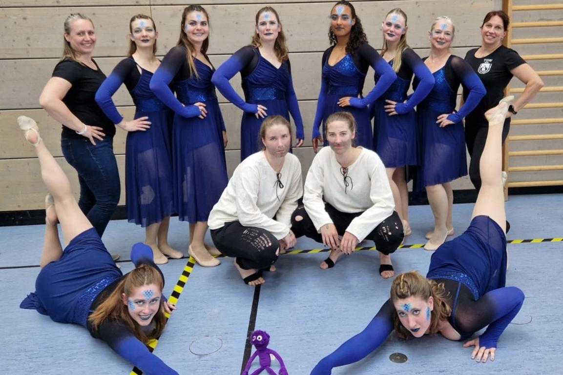 Gruppe von Tänzerinnen in blauen Kostümen posiert auf einer Sportfläche, einige in akrobatischen Posen, mit geschminkten Gesichtern.