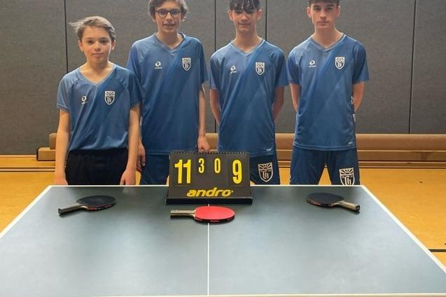 Vier Jugendliche in blauen Trikots stehen hinter einem Tischtennistisch mit einer Anzeigetafel (11:3, 0:9) und Schlägern.