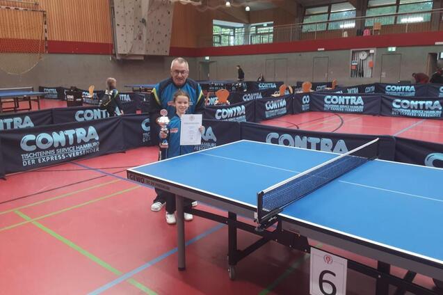 Ein Junge hält einen Pokal und ein Zertifikat in einer Sporthalle mit Tischtennisplatten im Hintergrund.