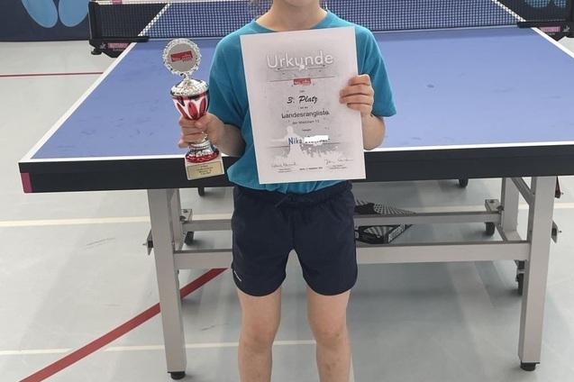 Mädchen hält einen Pokal und eine Urkunde für den 3. Platz in einem Tischtennisturnier, steht vor einer Tischtennisplatte.