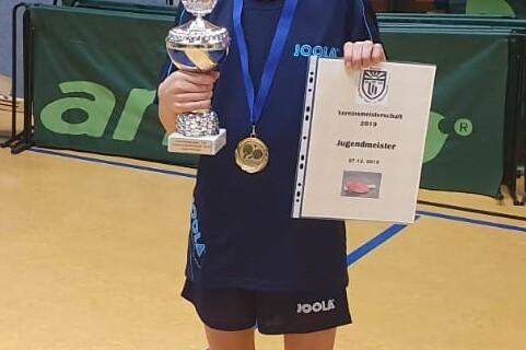 Junge mit Brille hält Pokal, Goldmedaille und Urkunde im Tischtennis in einer Sporthalle.