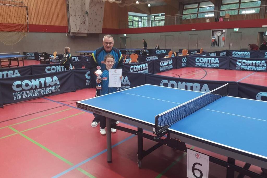 Ein Junge hält einen Pokal und ein Zertifikat in einer Sporthalle mit Tischtennisplatten im Hintergrund.