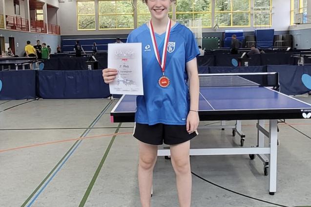 Junge Spielerin in blauer Trikot hält Urkunde und Medaille, steht vor einem Tischtennistisch in einer Sporthalle.