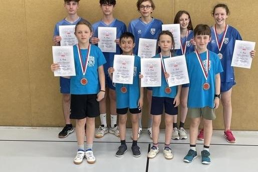 Gruppe von vier Jungen und fünf Mädchen in blauen T-Shirts mit Urkunden und Medaillen in einer Sporthalle.