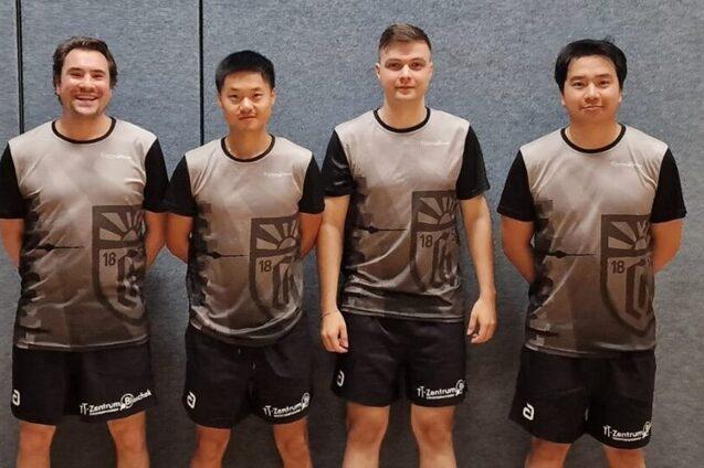 Vier Tischtennisspieler in grauen Teamtrikots posieren lächelnd vor einer grauen Wand in einer Sporthalle.