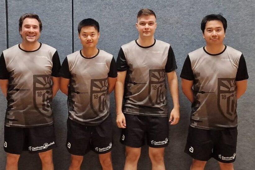 Vier Tischtennisspieler in grauen Teamtrikots posieren lächelnd vor einer grauen Wand in einer Sporthalle.