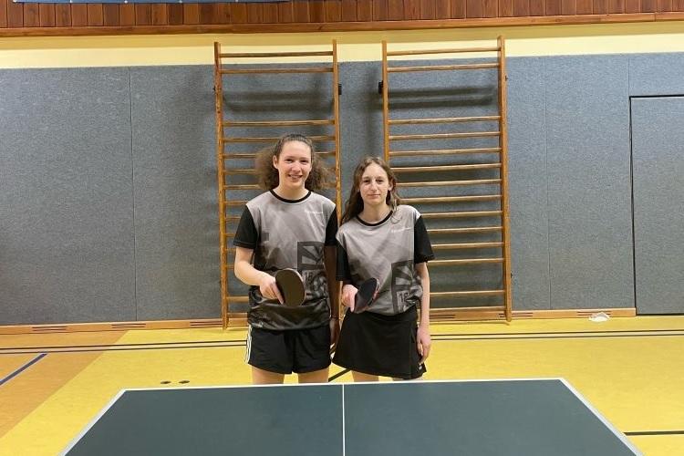 Zwei Tischtennisspielerinnen stehen an einem Tisch, bereit zum Spielen, in einer Sporthalle mit Banner im Hintergrund.