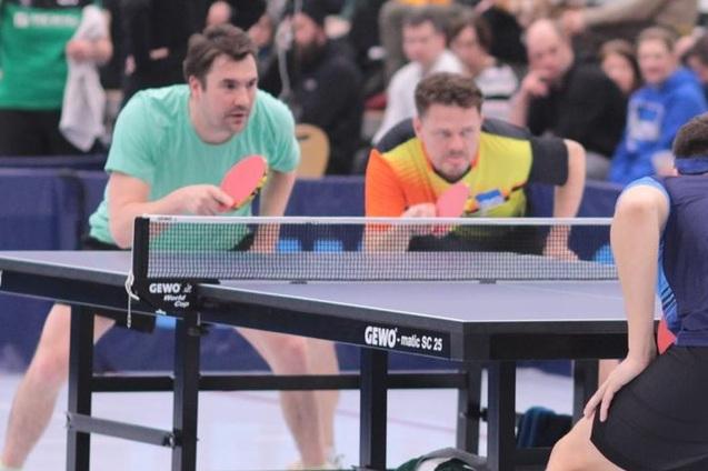 Zwei Männer in Sportkleidung hinter einem Tischtennistisch, bereit für den nächsten Ballwechsel. Zuschauende im Hintergrund.