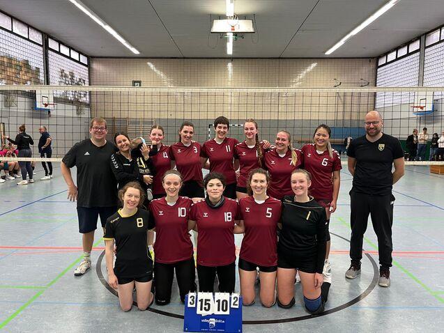 Gruppe von Frauenvolleyballspielern in roten Trikots, stehend und kniend vor einem Volleyballnetz in einer Sporthalle.