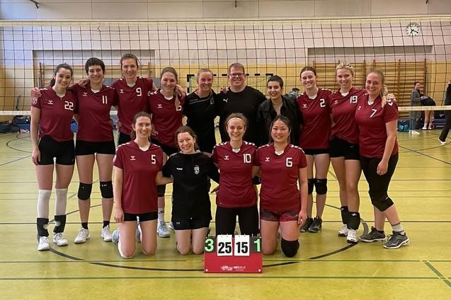 Mannschaftsfoto einer Volleyball-Truppe in roten Trikots, lächelnd vor einem Volleyballfeld mit Punktetafel.