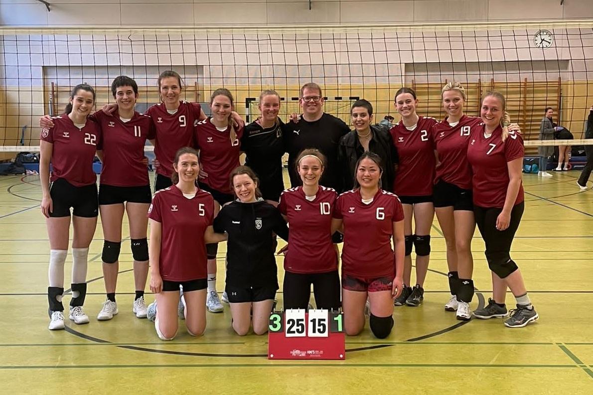 Mannschaftsfoto einer Volleyball-Truppe in roten Trikots, lächelnd vor einem Volleyballfeld mit Punktetafel.