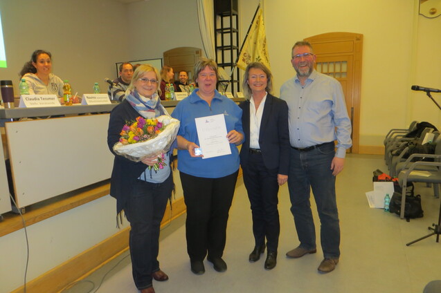 Vier Personen stehen auf einer Bühne, eine Frau hält einen Blumenstrauß und ein Zertifikat. Der Hintergrund zeigt einen Tisch mit Getränken.