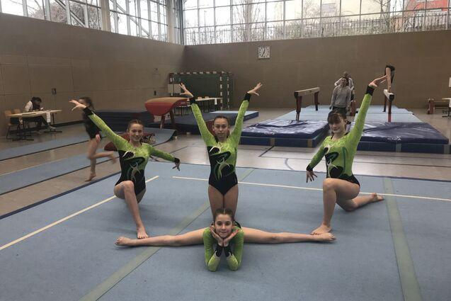 Vier junge Turnerinnen in grünen Bodenturnanzügen posieren auf einer Sportmatte in einer Turnhalle.