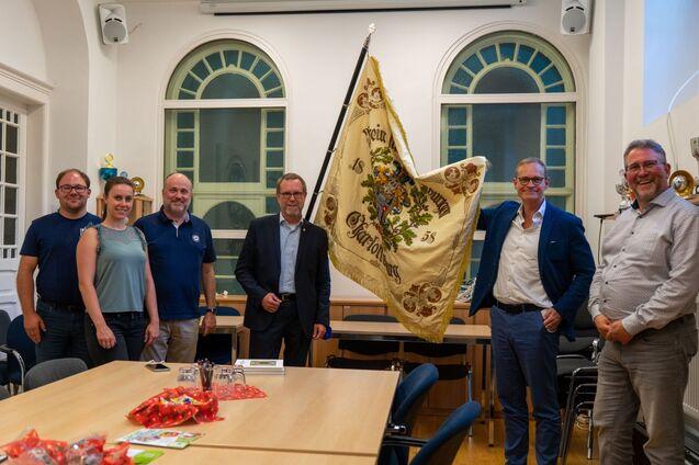 Gruppe von sechs Personen in einem Raum, eine Person hält eine Fahne mit Wappen, Tisch mit Getränken und Snacks im Vordergrund.