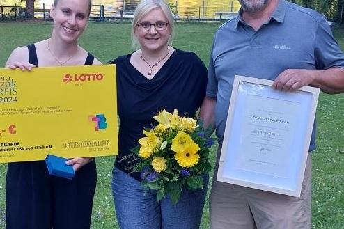 Drei Personen stehen auf einer Wiese, eine hält einen Scheck, die andere einen Blumenstrauß, alle lächeln. Im Hintergrund ein See.