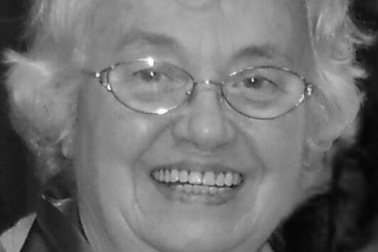 Lächelnde ältere Frau mit Brille und lockigem, grauem Haar in einem festlichen Outfit.