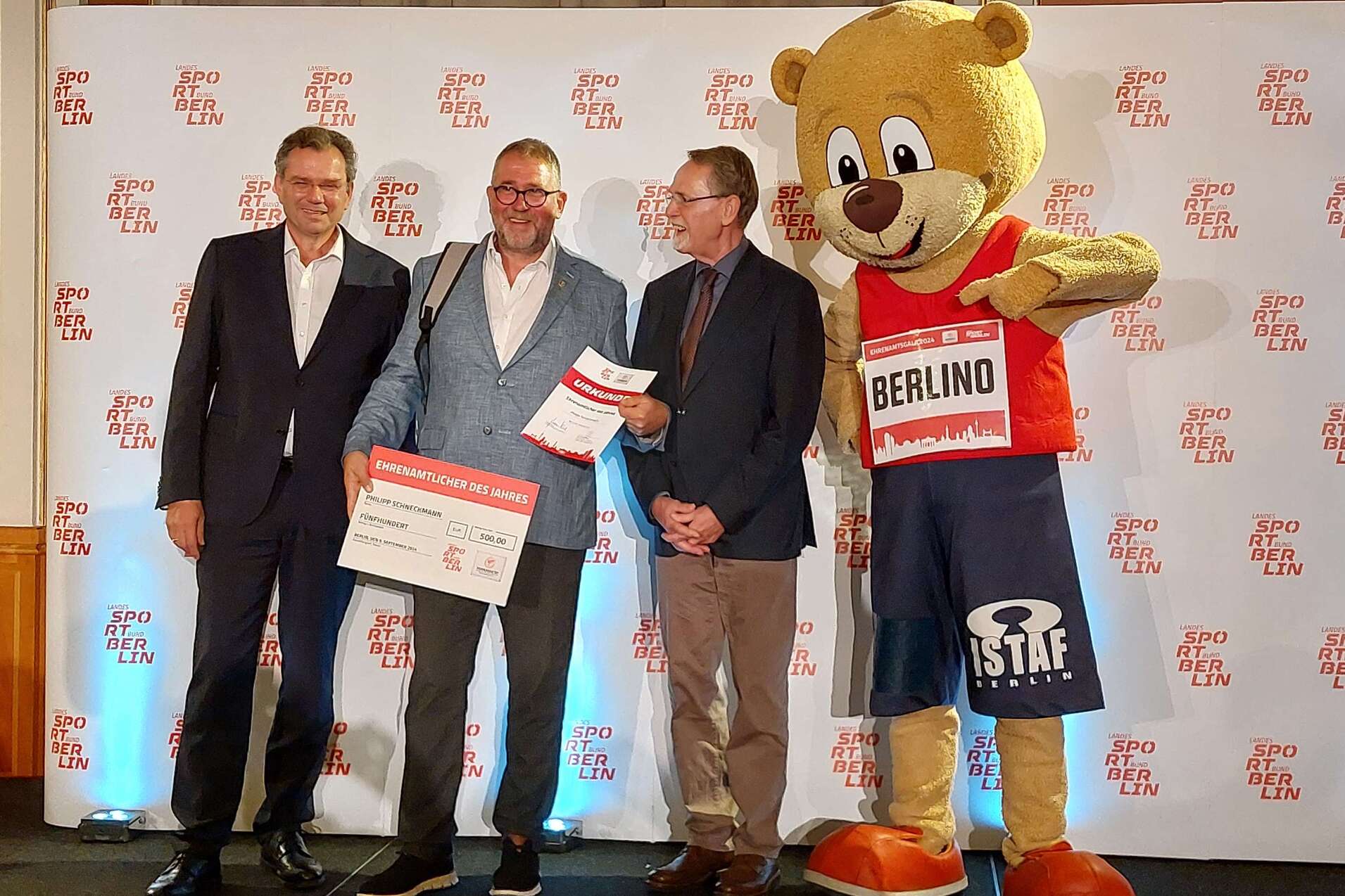 Drei Männer stehen auf einer Bühne, einer hält eine Urkunde, neben ihnen steht ein Bär in Sportkleidung.