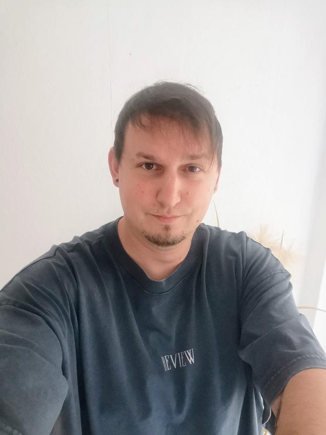 Nahaufnahme eines Mannes mit Kurzhaarfrisur, der ein graues T-Shirt mit Aufschrift trägt, vor einer weißen Wand.