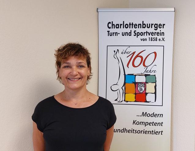 Frau mit Locken steht lächelnd vor einem Banner des Charlottenburger Turn- und Sportvereins mit Jubiläumslogo.