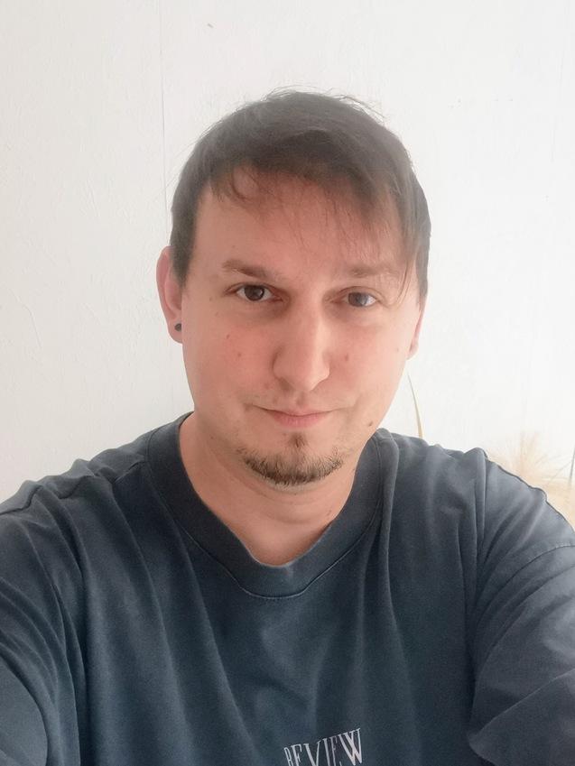 Nahaufnahme eines Mannes mit Kurzhaarfrisur, der ein graues T-Shirt mit Aufschrift trägt, vor einer weißen Wand.