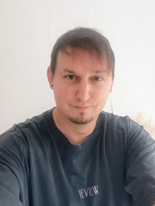 Nahaufnahme eines Mannes mit Kurzhaarfrisur, der ein graues T-Shirt mit Aufschrift trägt, vor einer weißen Wand.